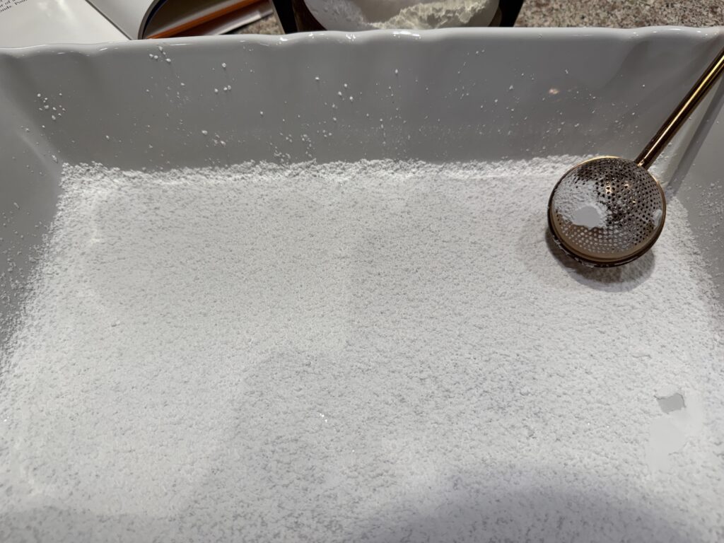 Rectangular baking pan being dusted with confectioners’ sugar using a flour duster for homemade marshmallows