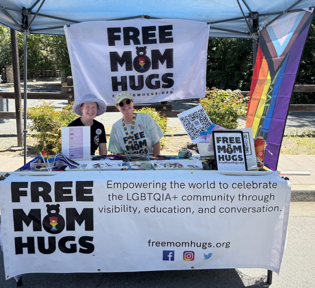 Free Mom Hugs booth at the Clayton Pride Festival with volunteers, flags, and information materials