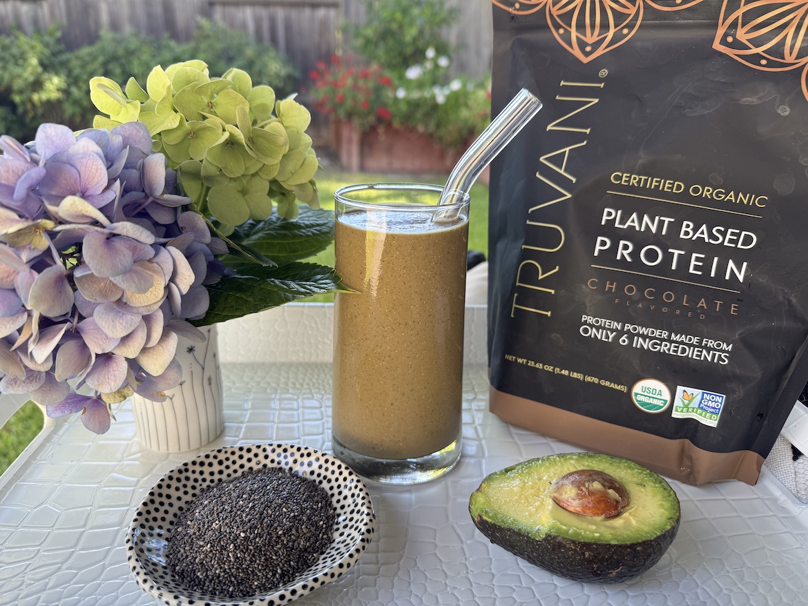 Deborah's Velvet Green Smoothie in a glass beside Truvani Chocolate Protein powder, topped with kiwi and chia seeds.