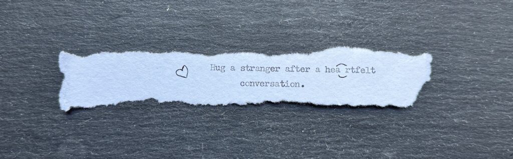 Torn typewritten note reading “Hug a stranger after a heartfelt conversation” with a hand-drawn heart
